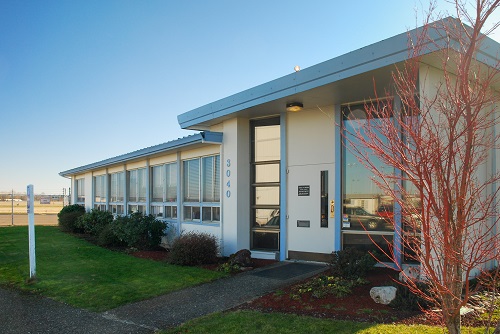 Single‑story mid‑century office building with light‑colored siding and a low‑sloped roof, situated on a paved lot adjacent to Salem Airport runways.