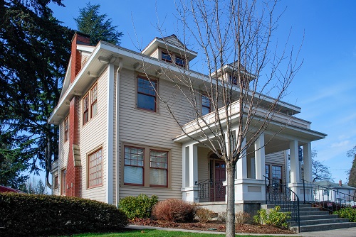 Two-story building with a light beige exterior and red-brick trim, featuring large windows—including prominent bay windows on the second floor—and a central columned porch.