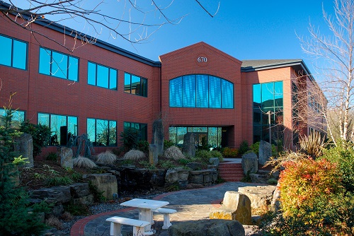 A two‑story mid‑century brick office building with large windows and a flat roof, set on a manicured lawn with trees.