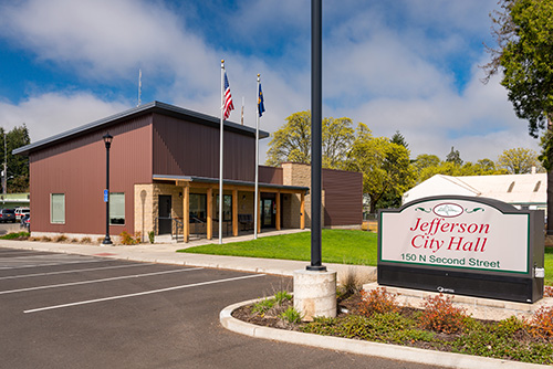 A modern single-story building with a brown and beige exterior. It features large windows and a flat roof. A sign in front says Jefferson City Hall.