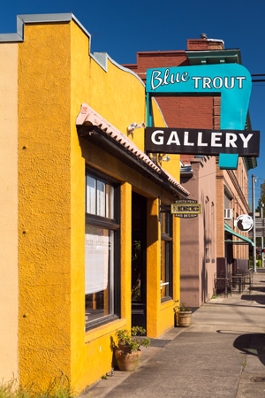 Yellow two-story Blue Trout Gallery building with a red‑tiled roof, large display windows, gallery sign above the entrance.