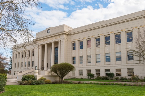 Front view of the Linn County Courthouse in Albany, Oregon: a classical white‑stone building with tall columns supporting a central portico, a clock set into the building’s pediment above the entry