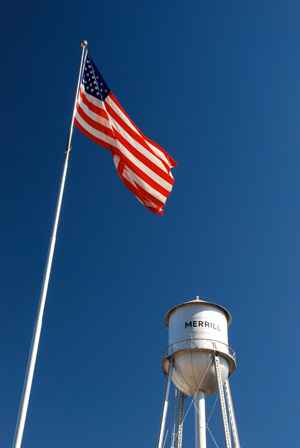 View of water tower with 'Merrill' printed on the side as seen from below. An american flag flies. Sky is blue with no clouds.