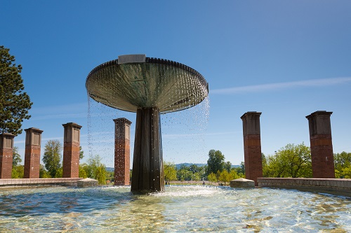 A circular stone fountain in a park in Independence, Oregon, featuring water cascading from a central column into a round basin.