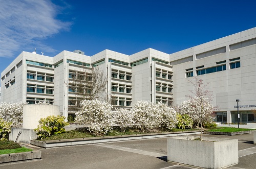 A mid‑height, rectangular government office building clad in light-colored brick or stone, with horizontal rows of windows. In the foreground are landscaped lawns, mature deciduous trees, and flowering shrubs.