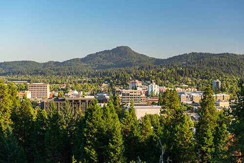 A panoramic view of a city nestled among lush green hills under a clear blue sky. The foreground is dominated by dense treetops, while the middle ground features various buildings, including mid-rise structures and possibly institutional facilities.