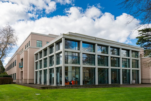 A mid‑rise, mid‑20th‑century office building with a rectangular footprint, clad in light-toned concrete or stone panels. The facade features evenly spaced vertical window strips across multiple floors. In front are manicured shrubs and small deciduous trees, bordered by a paved walkway.