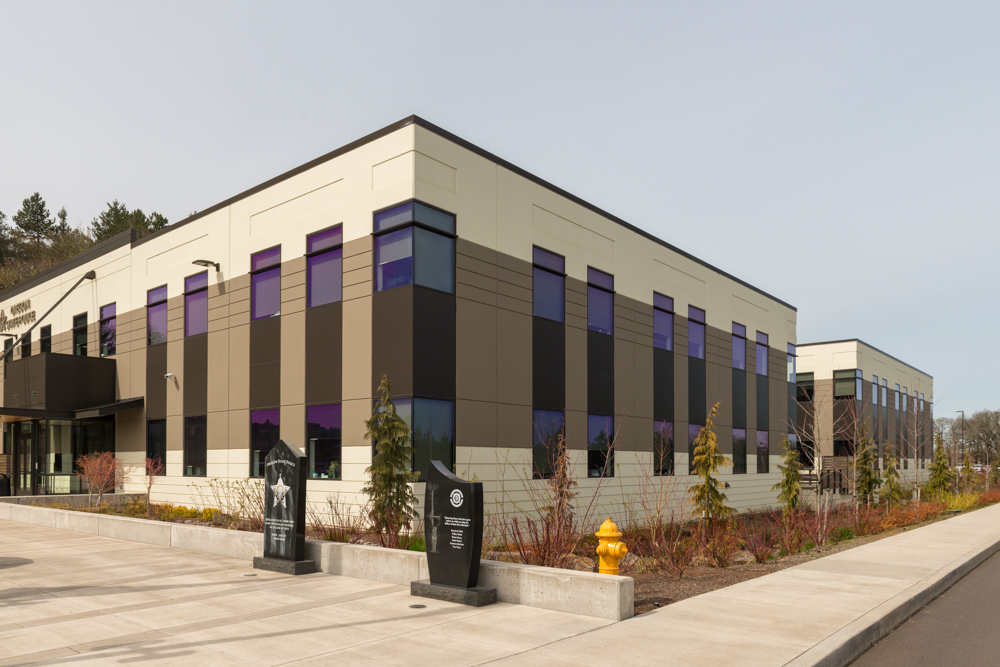 A modern, two-story government office building clad in light beige concrete or stucco, with large dark-framed windows spanning both floors. A flat roof tops the rectangular structure.
