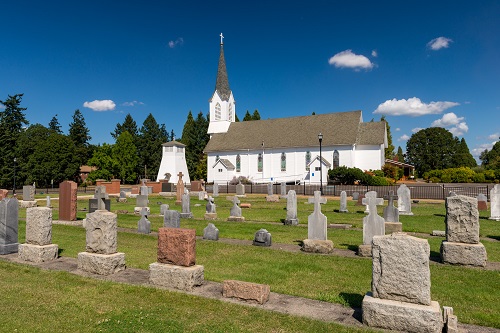 Single-story church with steeple in background. Foreground has neat rows of headstones for graves. Some have crosses for tops.