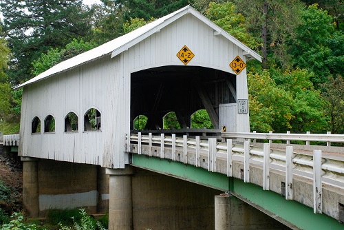 Covered bridge with side windows with curved tops. 4 windows on each side, portals with flat arched openings & exposed false beams at the gable ends.