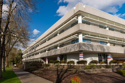 Veterans’ Building in Salem — a multi‑story, white‑faced office building with large rectangular windows and prominent red and blue columns at the entrance, surrounded by trees under a clear sky.