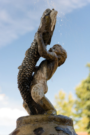 A stone fountain featuring a whimsical statue of a child holding a fish aloft, with water cascading from the fish’s mouth into a circular basin.