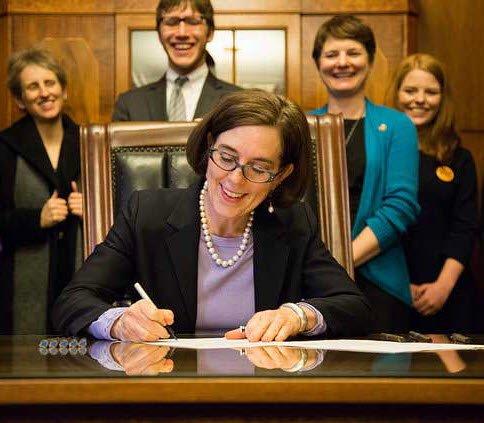 Kate Brown sitting at a desk signing the Oregon Motor Voter Law. Four people stand behind her smiling and watching her sign.
