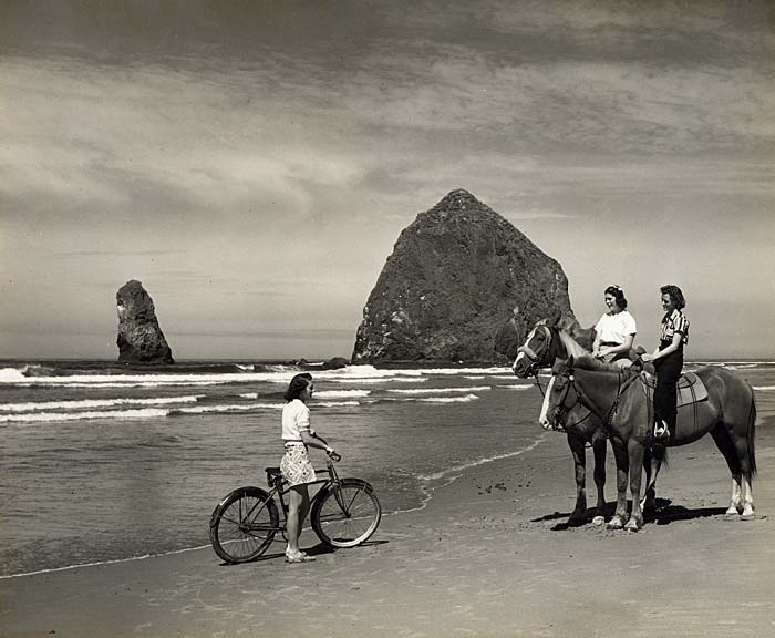 At Cannon Beach with Haystack rock in the back. A woman on bicycle talks with two women on horseback near water on the beach.