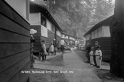 Women & children stand around a sidewalk lined with identical wood dwellings. 