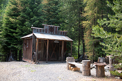 One room wood building with tin roof. Tall evergreen trees surround 2 sides of the structure. The other sides open to gravel.