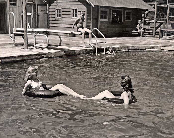 Two women float on rubber tubes in a pool. A boy sits on a diving board with feet hanging over side.