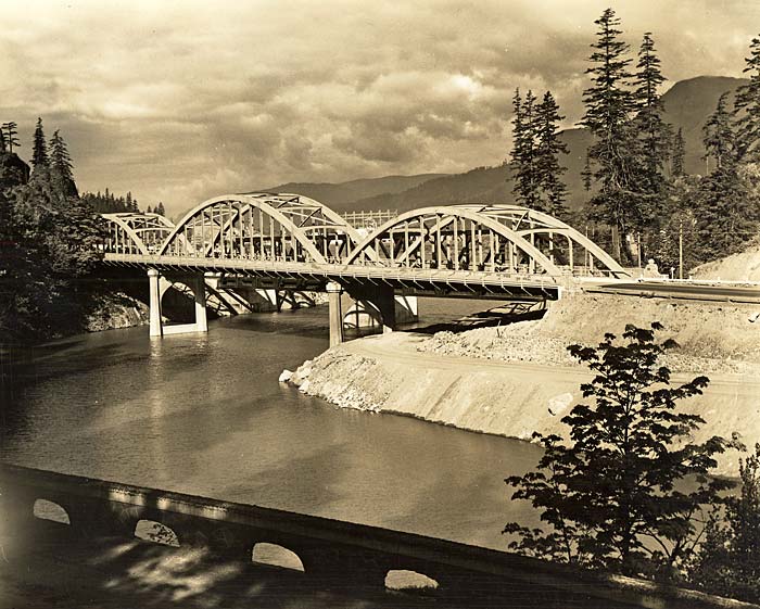 Eagle Creek Bridge spans a river with tall fir trees on each side and tall mountains in the background.