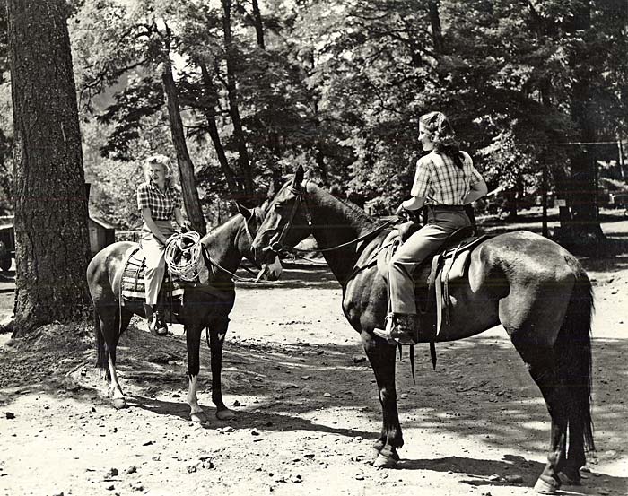 Two women sit on horseback in a forested area.