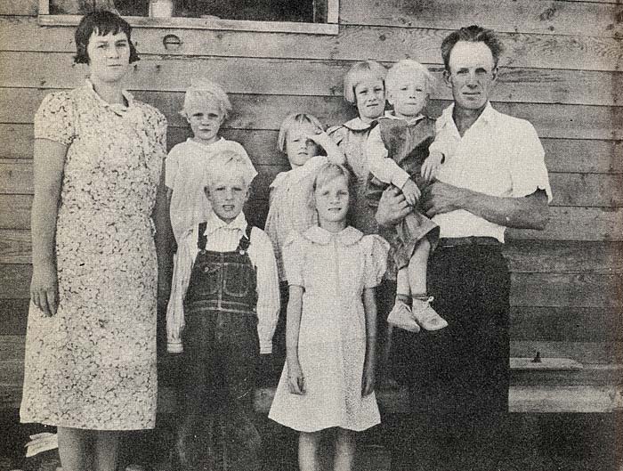 Family of 8 stand in front of wood plank house: mother, father and 6 children.