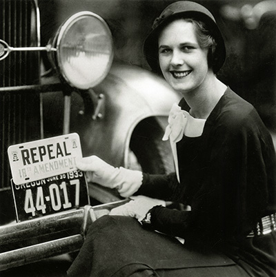 Stylish woman in white gloves and a cloche hat holds "Repeal 18th Amendment" license plate over a real license plate of a car.