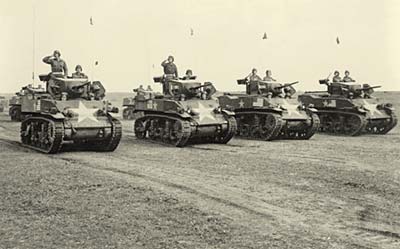 Line of 4 tanks with men atop.