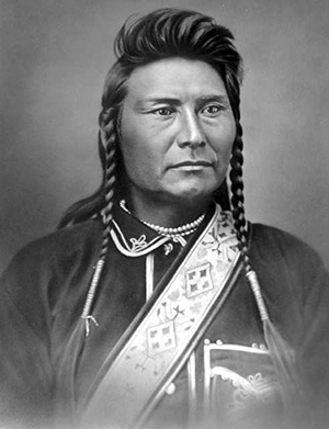 Picture of Chief Joseph wearing native clothing. His hair is short on top with 2 long braids coming down each side of the face. 