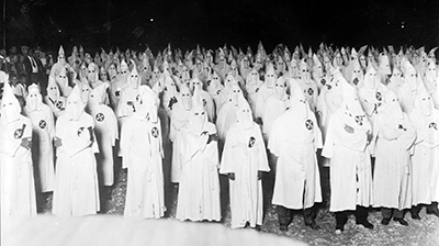 A hundred or more people dressed in KKK outfits with tall pointy hoods stand in a crowd.