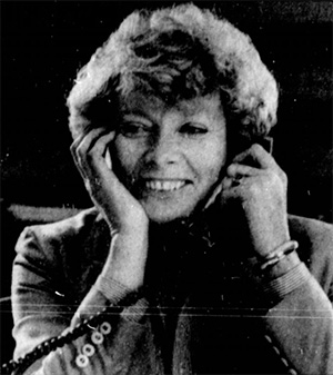 Norma Paulus holds a telephone receiver to one ear. She is smiling.