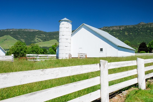 A barn with a grain silo attached at one end and a picket fencing running along the side. Low mountains are seen in the back.