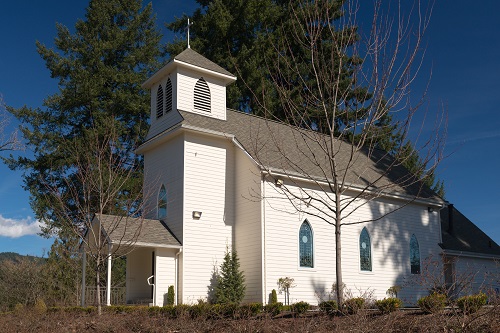 Small church with bell town in front topped with cross. Stained glass windows line the side. Front porch is covered.