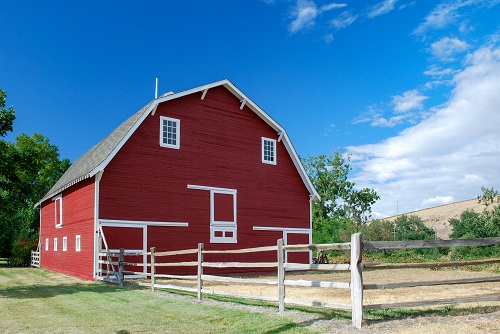 Traditional red barn with wooden fence surrounding a corral area attached. Mowed green lawn extends out from the corral.