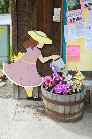 Colorful flowers in a round wood planter. A painted wood representation of a girl watering the flowers.