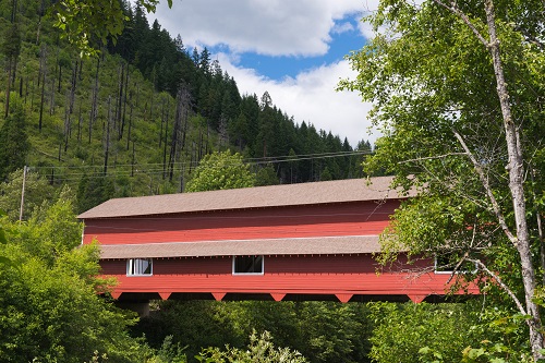 180 foot covered bridge using triple Howe Truss construction. A mix of evergreen & deciduous trees surround.